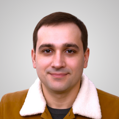 A man with short dark hair wearing a brown jacket with a white collar.